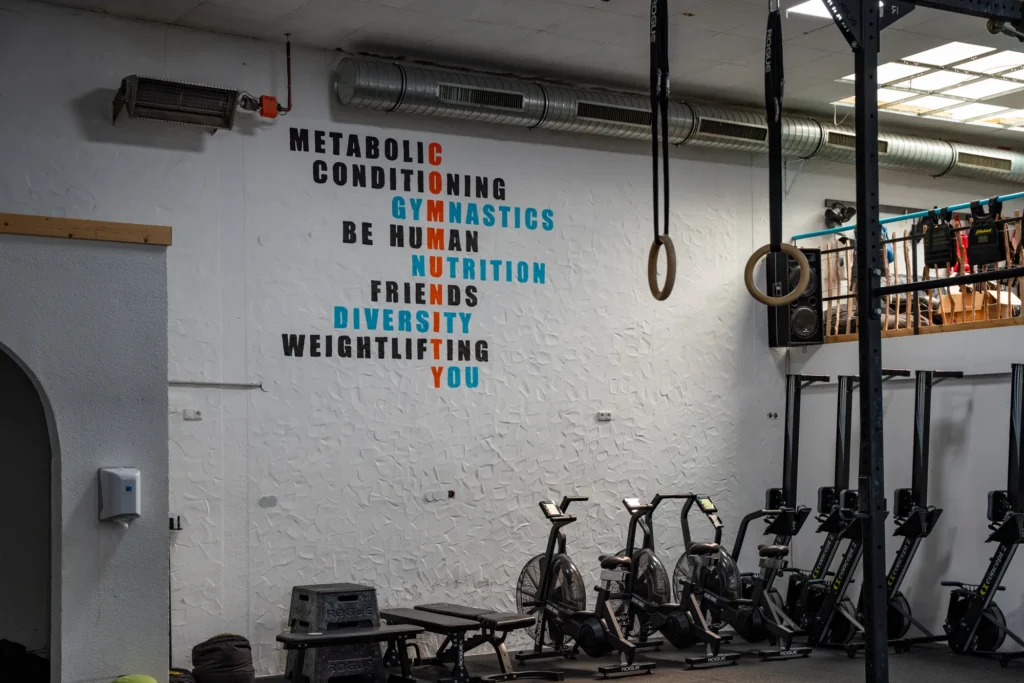 Hallenwand mit dem Schriftzug Metabolic Conditioning, Gymnastics, Be Human, Nutrition, Friends, Diversity, Weightlifting, You; davor Rudergeräte und Bikes