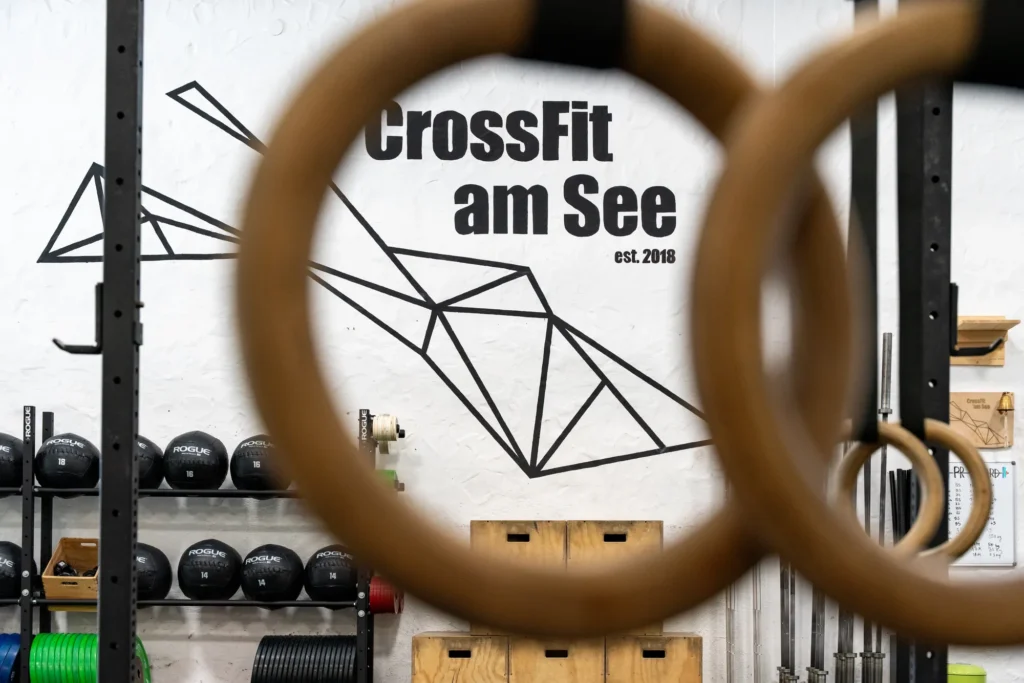 Unscharfe Turnringe im Vordergrund, dahinter das Logo von CrossFit am See an der weißen Hallenwand