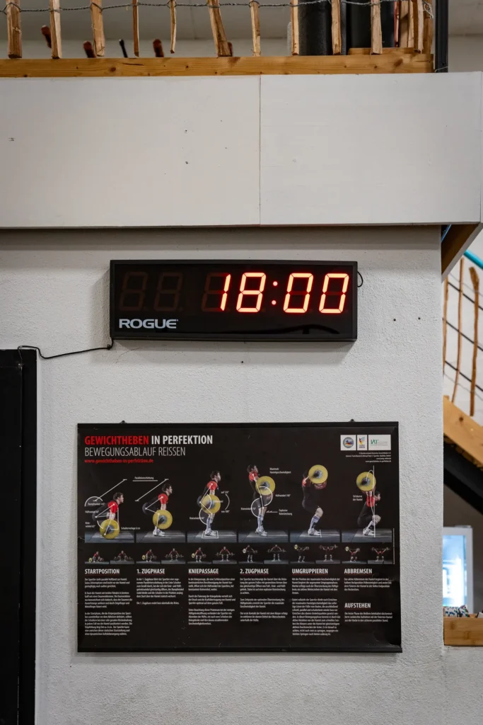 Digitale Trainingsuhr mit der Anzeige 18:00 über einer Übungstafel zum Gewichtheben